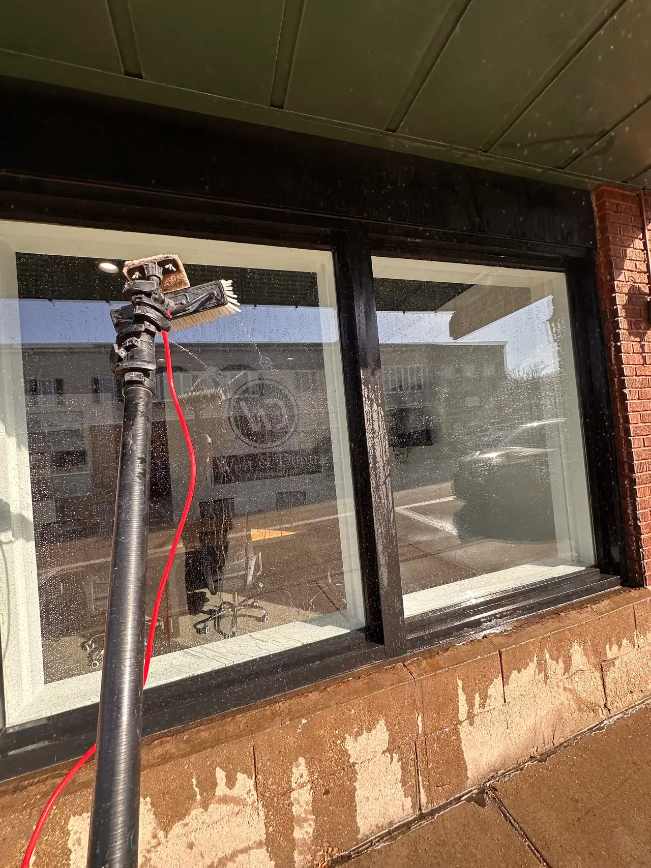 A water-fed pole brush cleaning a storefront window, red pure-water hose and brick sill — Madison, WI