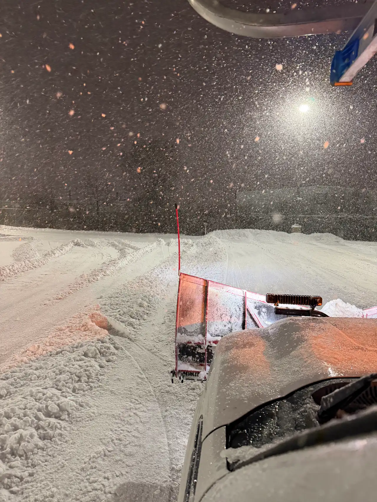 A red snow plow blade pushing snow across a lot at night, seen from the truck cab — Madison, WI