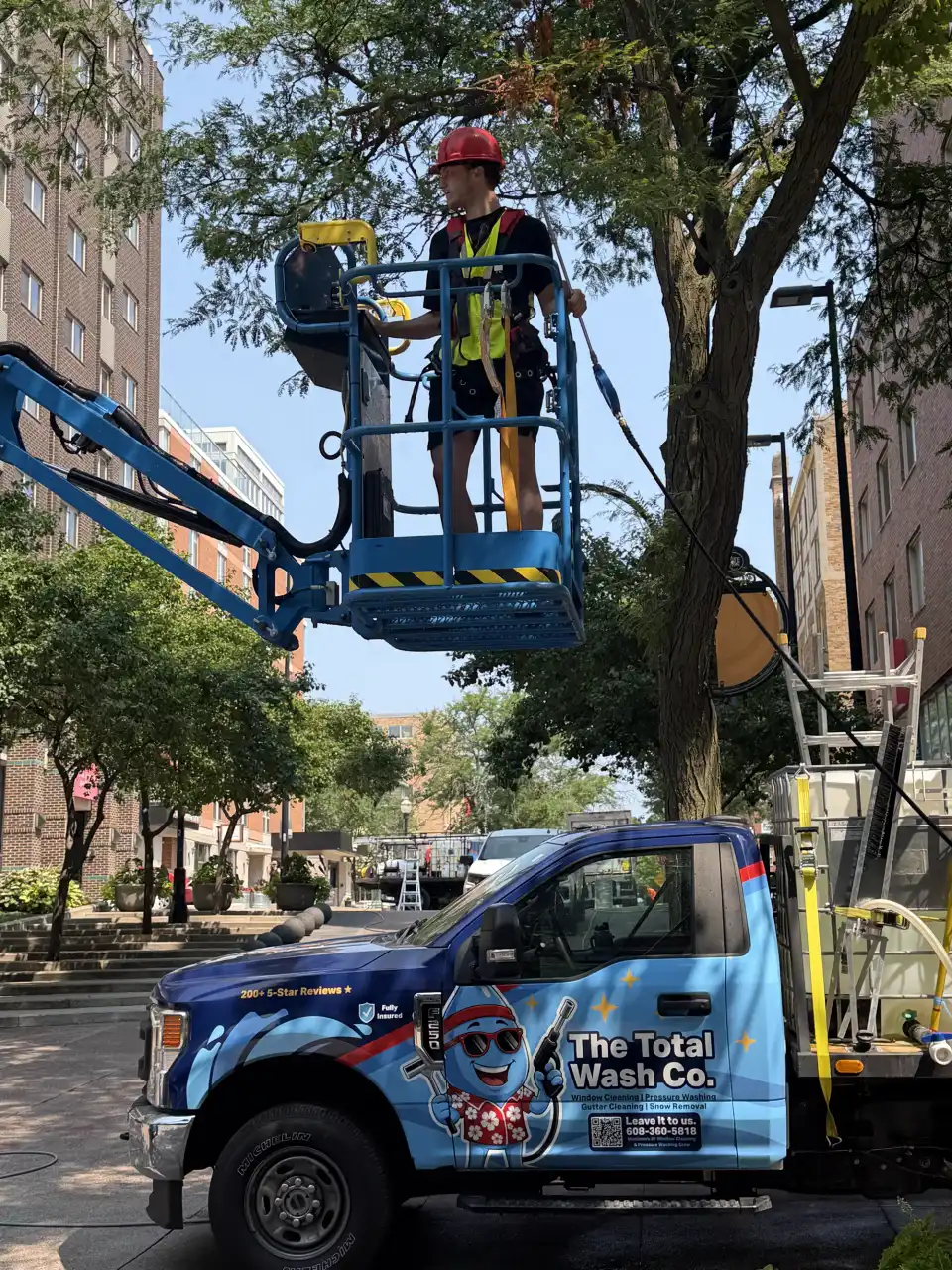 A Total Wash Co. technician in a lift basket at the upper-floor windows of a brick building, the company truck parked below